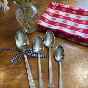 May include: Four silver-toned metal spoons of varying sizes arranged on a wooden surface. The largest spoon is tied with a navy and white checkered ribbon. A clear glass vase with yellow flowers and a red and white patterned cloth are nearby.