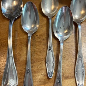 May include: A set of five silver-toned metal spoons arranged in a row. The spoons feature a polished finish with decorative embossed floral patterns on the handles. The leftmost spoon has a distinct flared base, while the others have tapered handles.