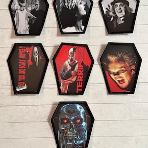 Może przedstawiać: Kolekcja siedmiu naszywek w kształcie trumny z postaciami i obrazami z klasycznych horrorów. Naszywki przedstawiają obrazy Frankensteina, narzeczonej Frankensteina i innych kultowych postaci. Niektóre naszywki mają tekst "DO YOU LIKE SCARY MOVIES?" i "TERRIFIER".