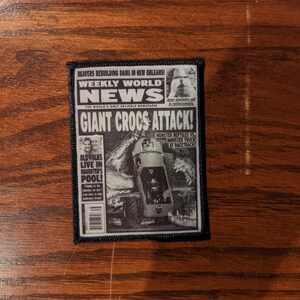 May include: A black and white patch featuring a newspaper design with the headline "GIANT CROCS ATTACK!". The patch includes the text "WEEKLY WORLD NEWS" and other news stories. The patch is rectangular with a black border.