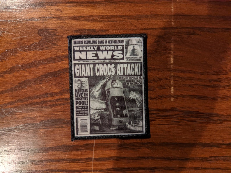 May include: A black and white patch featuring a newspaper design with the headline "GIANT CROCS ATTACK!". The patch includes the text "WEEKLY WORLD NEWS" and other news stories. The patch is rectangular with a black border.