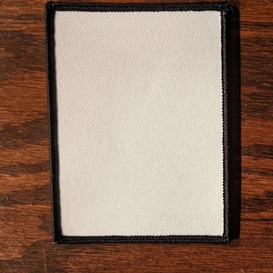 May include: A rectangular white patch with a black border, on a wooden surface. The patch has a textured surface and is framed by a black stitched edge. The background is a dark brown wood grain.