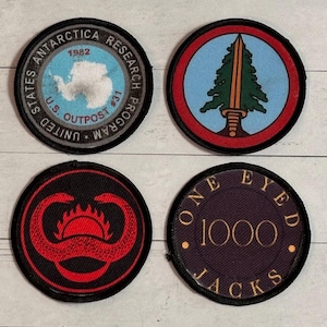 Könnte beinhalten: Vier runde, gestickte Aufnäher. Einer zeigt eine Karte der Antarktis mit dem Text "U.S. Outpost #31" und "United States Antarctica Research Program 1982". Andere Aufnäher zeigen einen Baum und ein Schwert, ein rotes Design auf Schwarz und "One Eyed 1000 Jacks".