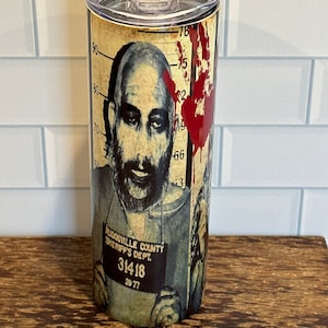 May include: A stainless steel tumbler featuring a mugshot design with a man's face, blood-red handprint, and the text "PLIGIOSVILLE COUNTY SHERIFF'S DEPT. 31418 29 77". The tumbler has a clear lid.