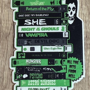 Puede incluir: Una pegatina con una pila de cintas VHS con títulos como "The Misfits" y "Astro Zombies". El diseño incluye una combinación de colores verde y negro, un gráfico de calavera y el texto "All the Hits From Hell".
