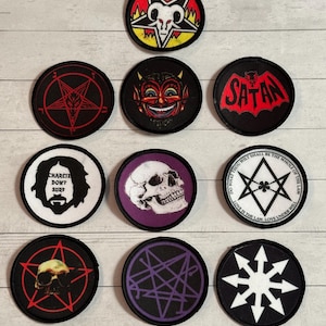 May include: A collection of round, embroidered patches with various occult and gothic designs. The patches feature images of a skull, a devil, a goat head, and pentagrams. Some patches have text, including "Satan" and "Charlie Don't Surf."