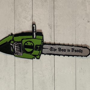 Puede incluir: Parche bordado con un diseño de motosierra verde y negro. El parche incluye el texto "The Saw is Family" en fuente gótica, con una hoja plateada. Ideal para ropa o accesorios.