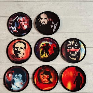 May include: A collection of nine circular patches featuring various horror movie and music icons. The patches display images of characters and text, with a variety of colors including red, black, and white. The patches are arranged on a light wood-grain surface.