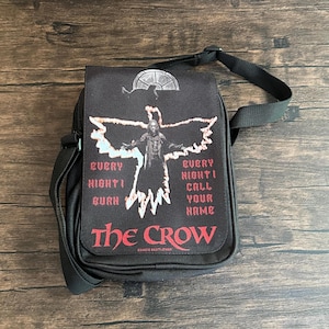 Könnte beinhalten: Schwarze Umhängetasche mit einer Grafik von "The Crow". Das Design zeigt eine Figur mit ausgestreckten Armen, den Text "Every night I burn, every night I call your name" und den Titel "The Crow".