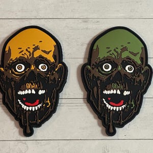 May include: Two zombie head patches. One patch has a yellow and brown head with white teeth and a red mouth. The other patch has a green and brown head with white teeth and a red mouth. Both patches have a black border.