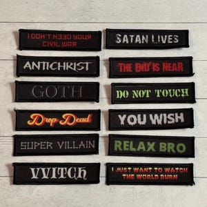 May include: A selection of black embroidered patches, each bearing a different phrase in white, red, or green lettering. Phrases include "Satan Lives," "Antichrist," "Goth," "Drop Dead," "Super Villain," "Witch," and others. The patches are rectangular.