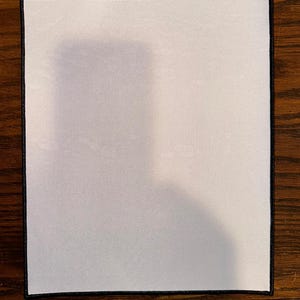 May include: A rectangular white sublimation blank with a black stitched border, resting on a wooden surface. The blank is designed for custom printing or heat transfer projects. The image shows the front of the blank.