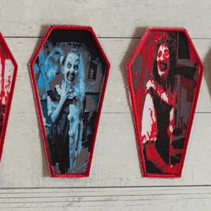 May include: Four coffin-shaped patches with horror-themed designs. Three have red borders, and one has a green border. The images inside depict scary figures, with one in blue tones. The patches are likely for clothing or accessories.