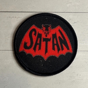 May include: A round, black patch with a red bat-like design and the word "SATAN" in bold, black letters. The patch has a stitched border and is likely made of fabric. The design is centered on the patch.
