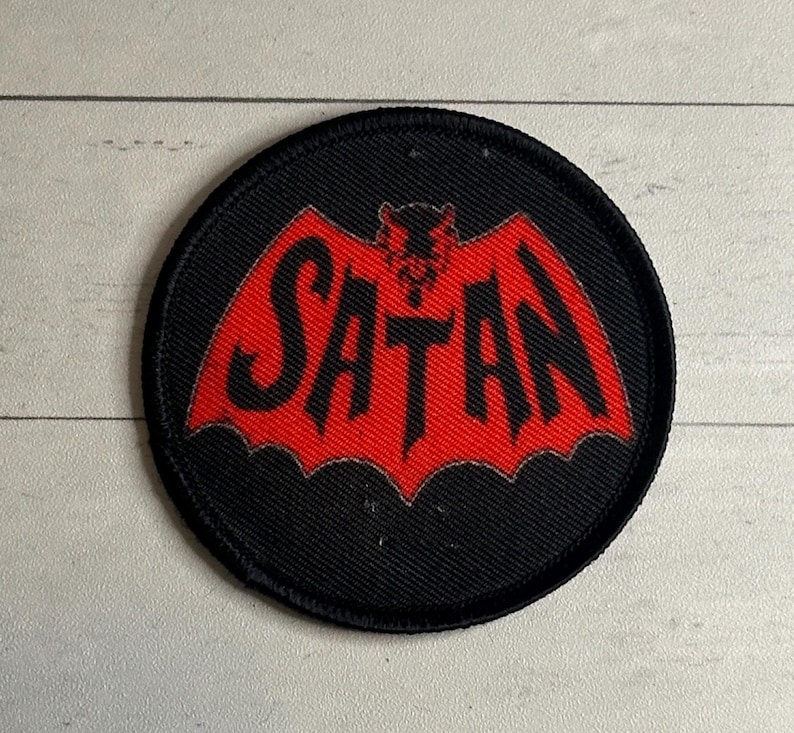 May include: A round, black patch with a red bat-like design and the word "SATAN" in bold, black letters. The patch has a stitched border and is likely made of fabric. The design is centered on the patch.