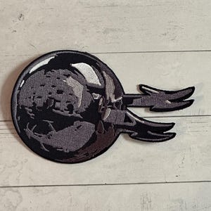 May include: A black and gray embroidered patch featuring a circular design with a detailed, textured pattern. Two stylized, wing-like shapes extend from the circle. The patch is likely for clothing or accessories.