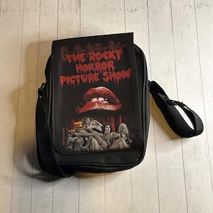 May include: Black messenger bag featuring the text "The Rocky Horror Picture Show" in red, with a red lip graphic. The bag has a black shoulder strap and a flap closure. The image on the bag also includes a group of people.