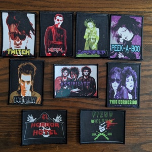 以下が含まれることがあります： 様々な画像とテキストが描かれた、黒い長方形のパッチのコレクションです。「TWITCH」、「METAL」、「ZOMBIFIED」、「HORROR HOTEL」などのポートレート、バンド名、フレーズが含まれています。これらのパッチは、おそらく衣類やアクセサリー用です。