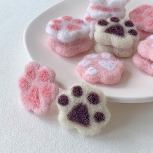 May include: A collection of felt paw print ornaments in pink, white, and brown. The paw prints are stacked on top of each other and are arranged on a white plate.