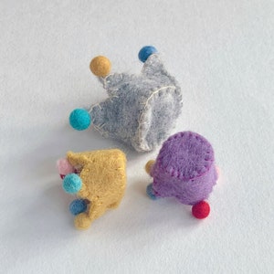 May include: Three small, handmade felt crowns in gray, yellow, and purple. Each crown features visible white or yellow stitching along the edges and is decorated with small, colorful felt balls in shades of blue, red, and pink attached to the points.