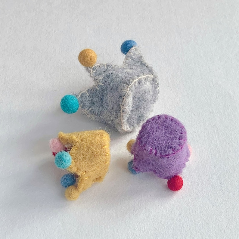 May include: Three small, handmade felt crowns in gray, yellow, and purple. Each crown features visible white or yellow stitching along the edges and is decorated with small, colorful felt balls in shades of blue, red, and pink attached to the points.