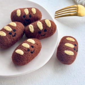 May include: Five small, brown, needle-felted bread rolls with cream-coloured slash details on top. Each roll features a simple, hand-stitched black thread face with two eyes and a curved smile. The felted items are arranged on a white plate.