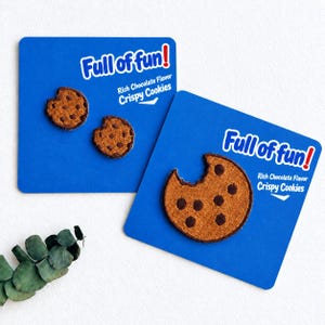 Pode incluir: Três emblemas bordados de bolachas com pepitas de chocolate fixados em cartões de apresentação azuis. Cada emblema é castanho com pepitas circulares castanho-escuras e uma marca de dentada. Os cartões apresentam o texto branco Full of fun! Rich Chocolate Flavor Crispy Cookies.