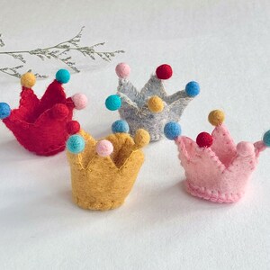May include: Four small felt crowns in red, yellow, light pink, and light gray. Each crown features a jagged top edge with small, colorful felt balls attached to the points in shades of blue, yellow, pink, and red, stitched with visible thread.