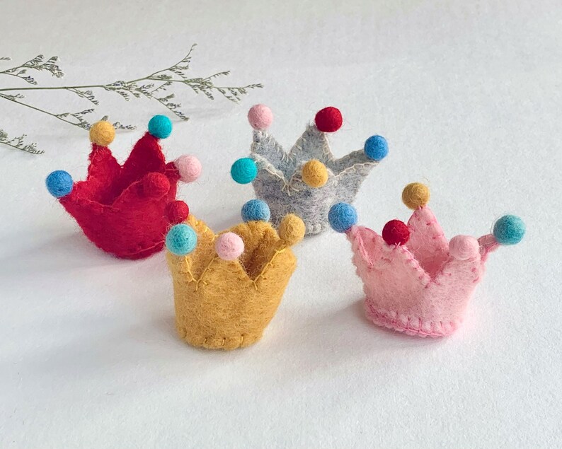 May include: Four small felt crowns in red, yellow, light pink, and light gray. Each crown features a jagged top edge with small, colorful felt balls attached to the points in shades of blue, yellow, pink, and red, stitched with visible thread.