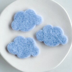 May include: Three light blue felt clouds on a white plate. The clouds are handmade and have a soft, fuzzy texture.