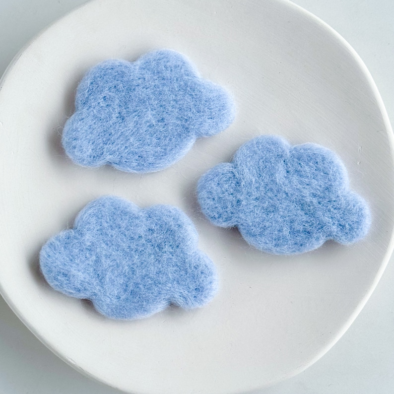 May include: Three light blue felt clouds on a white plate. The clouds are handmade and have a soft, fuzzy texture.