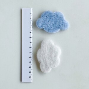 May include: Two felt clouds, one light blue and one white, are shown next to a ruler. The clouds are approximately 4 inches wide.