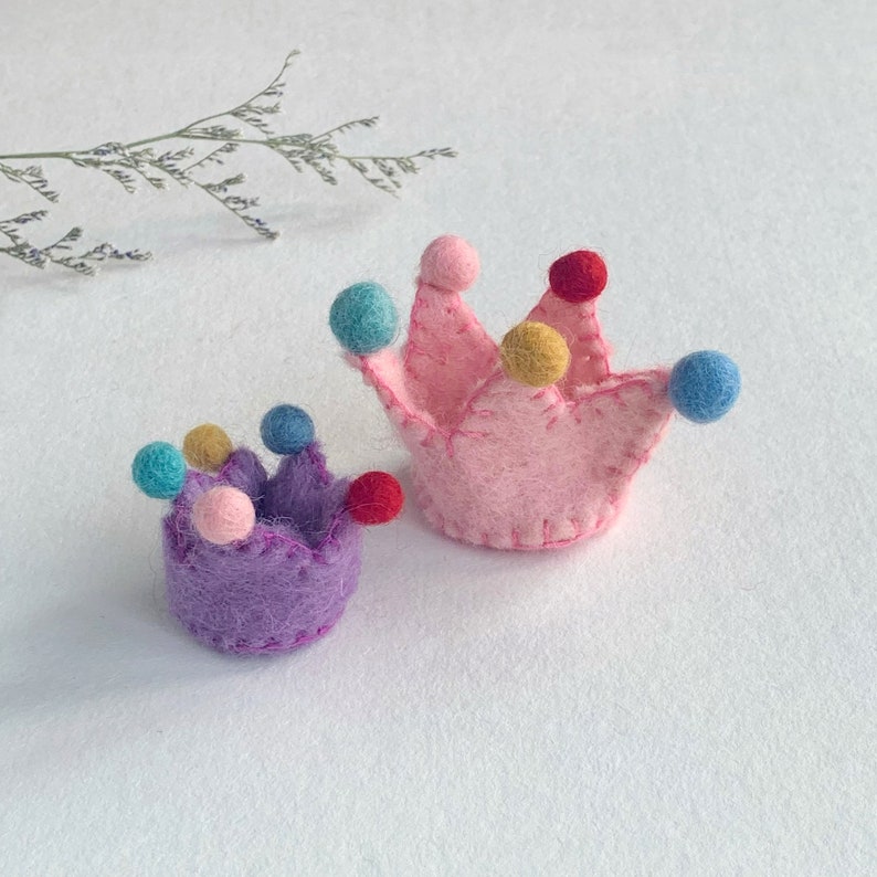 May include: Two felt crowns, one larger pink crown and one smaller purple crown. Each crown features stitched edges and is decorated with small, colorful felt pom-poms in shades of blue, yellow, pink, and red attached to the pointed tips.