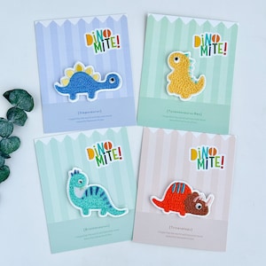 May include: Four colourful iron-on patches featuring different dinosaurs with the text "Dino Mite!" on each patch. The dinosaurs are a Stegosaurus, Tyrannosaurus Rex, Brontosaurus, and Triceratops.
