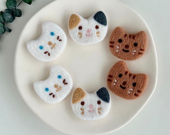 May include: Five felt cat brooches in various colors and designs. The brooches are white, brown, and black with embroidered details.