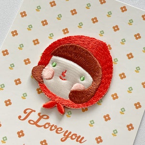 以下が含まれることがあります： 赤いフード付きのマントを着た茶色の髪の漫画の女の子が描かれた刺繍のアイロン接着パッチ。パッチはオレンジと緑の花が繰り返し描かれた白い背景の上にあります。「I Love you」というテキストがパッチの下に書かれており、その下に「As I have loved you, so you must love one another.」というテキストが続きます。