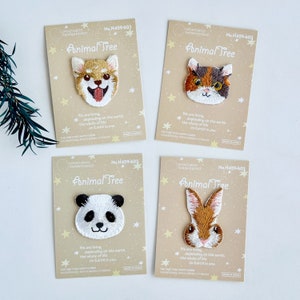 Könnte beinhalten: Vier gestickte Bügelbilder mit Tiergesichtern: ein Shiba Inu, eine dreifarbige Katze, ein Panda und ein braunes Kaninchen. Jedes Bügelbild befindet sich auf einer beige Karte mit dem Text "Animal Tree" und "We are living, depending on the earth. The whole of life on Earth is one."
