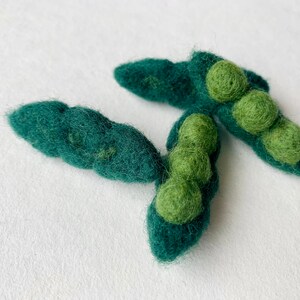 May include: Three felt green pea pods with green felt peas inside. The pea pods are arranged in a cluster on a white background.