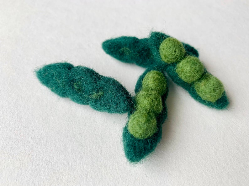May include: Three felt green pea pods with green felt peas inside. The pea pods are arranged in a cluster on a white background.