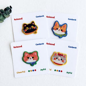 May include: Four embroidered cat patches on white cards. One is a black cat with yellow trim, one is an orange and white tabby with a green scarf, one is a red and white tabby with a blue scarf, and one is a grey cat with a red bow and collar.