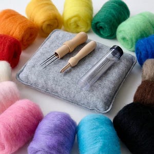 Op de afbeelding: Een knutselset met kleurrijke wol in rood, oranje, geel, groen, blauw, roze, paars, bruin, zwart en wit. Inclusief een grijs viltkussen, houten naaldgereedschap en een buisje naalden voor naaldvilten.