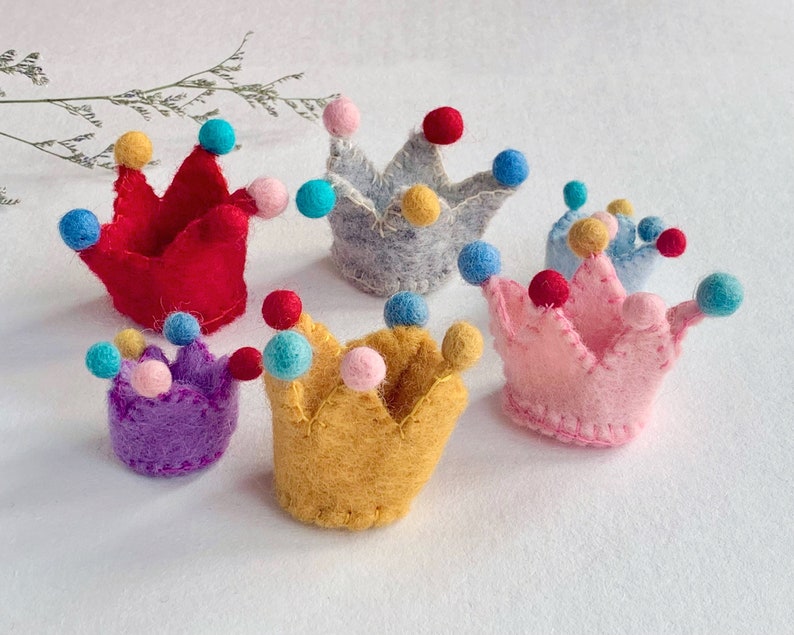 May include: A collection of six small felt crowns in various colors including red, gray, yellow, purple, pink, and light blue. Each crown features stitched edges and is decorated with small, colorful felt pom-poms attached to the points.