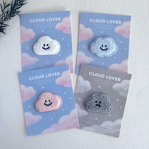 Könnte beinhalten: Vier Karten mit wolkenförmigen Aufnähern. Jede Karte hat einen unterschiedlich farbigen Wolkenaufnäher: weiß, hellblau, rosa und grau. Die Karten haben einen blauen oder grauen Hintergrund mit Wolken- und Sternillustrationen. Der Text "CLOUD LOVER" ist auf jeder Karte aufgedruckt.