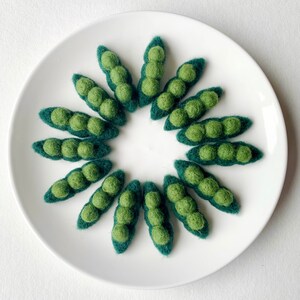 May include: A circle of felt pea pods in shades of green arranged on a white plate. The pea pods are made of felt and have a realistic appearance.