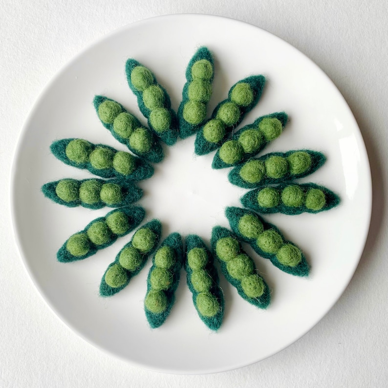 May include: A circle of felt pea pods in shades of green arranged on a white plate. The pea pods are made of felt and have a realistic appearance.