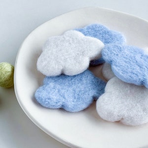 May include: A white plate with four felt cloud shapes in shades of blue and white. The clouds are arranged in a pile, with the white clouds on top.
