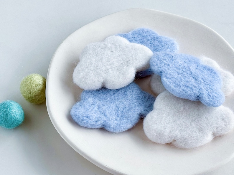 May include: A white plate with four felt cloud shapes in shades of blue and white. The clouds are arranged in a pile, with the white clouds on top.