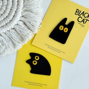 May include: Two black cat embroidery patches with yellow eyes rest on yellow cards. The cards have the text "BLACK CAT EMBROIDERY PATCH" and "I hope that the world will become more beautiful because of you."