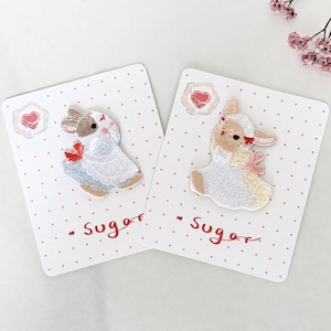 May include: Two embroidered rabbit patches on white cards with red polka dots and the word Sugar. One rabbit is brown and white with a red bow, the other is tan and white with a pink bow. Both wear white aprons and are displayed on individual cards.