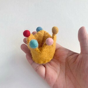May include: A small, mustard yellow felt crown featuring five points, each topped with a tiny, round, felted ball in red, light blue, dark blue, pink, and tan. The crown is constructed with visible stitching along the edges and base.